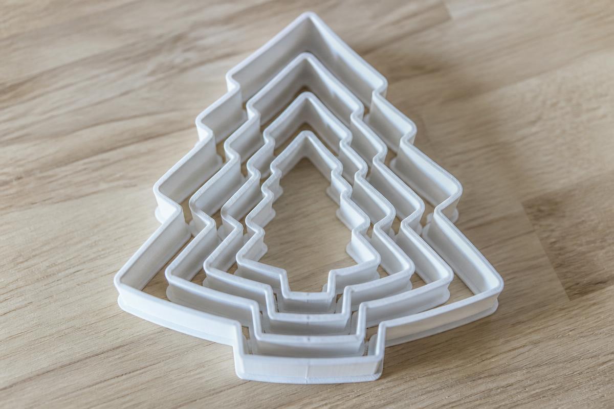 A nested set of five white 3D printed Christmas tree cookie cutters in graduated sizes, stacked one inside the other on a timber bench