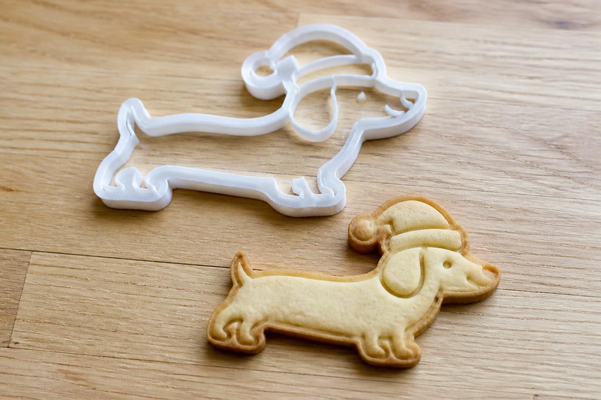 A custom white 3D printed cookie cutter shaped like a dachshund in a santa hat, next to a baked cookie of the same shape with its details pressed in