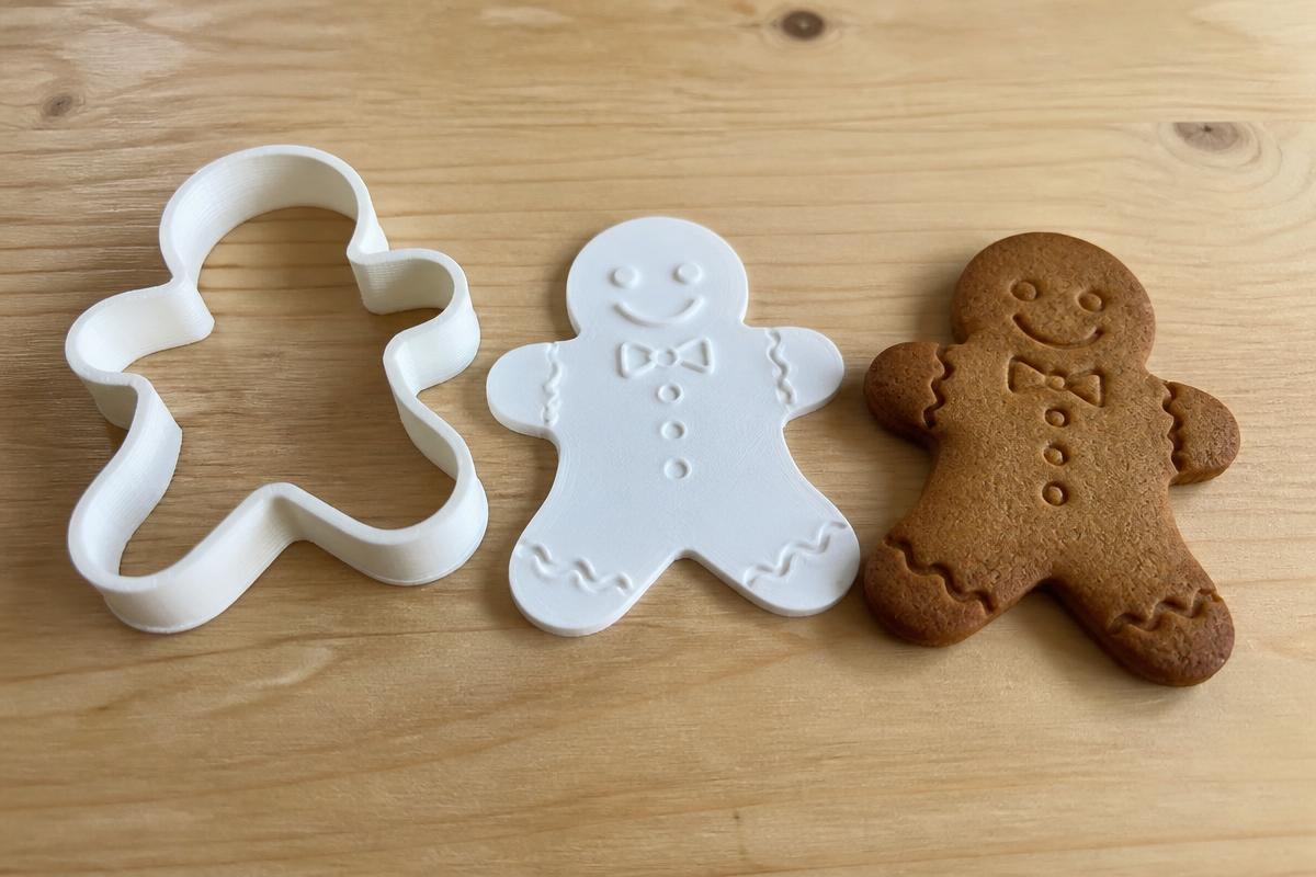 A white 3D printed gingerbread man cookie cutter beside its matching detail stamp plate and a baked gingerbread cookie with a smile, buttons and a bow pressed into the surface